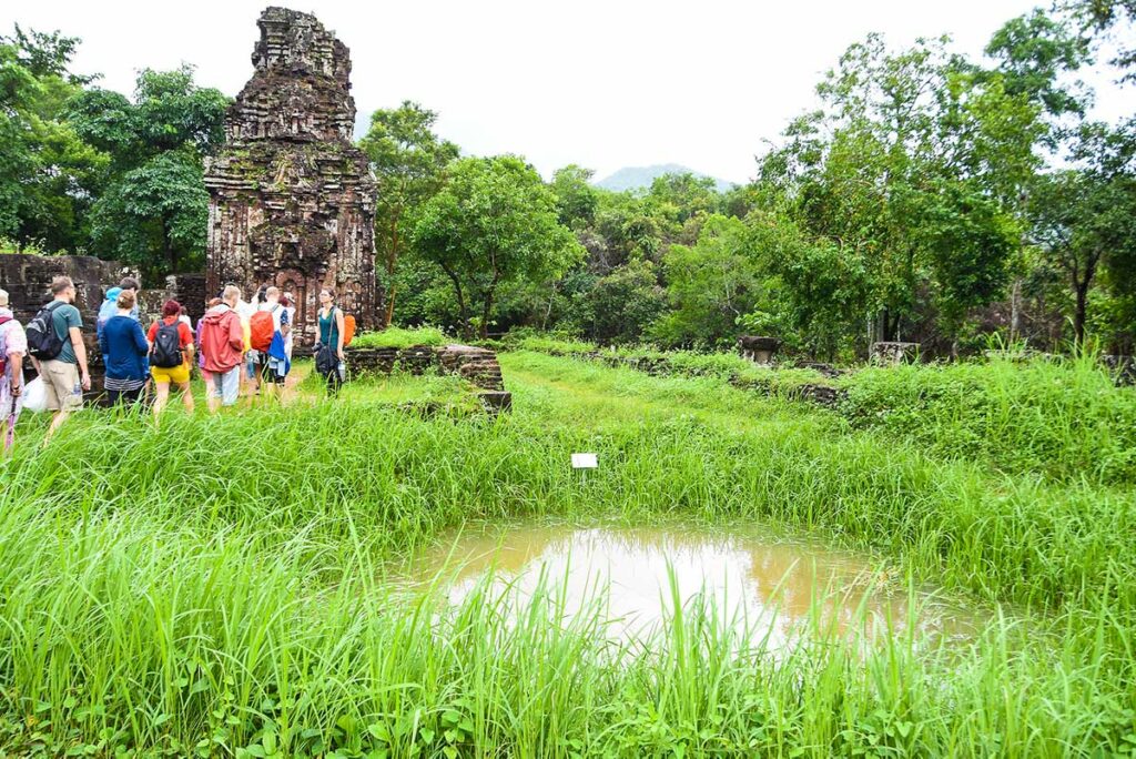 Group of visitors walking past a war-era bomb crater at My Son Sanctuary, a visible reminder of the damage the site suffered during the Vietnam War