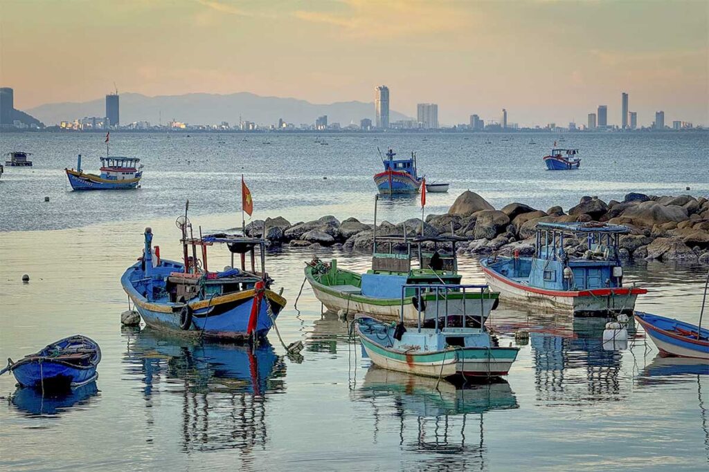 Fishing boats at Bai Xep Fishing Village with Quy Nhon city skyline – Colorful wooden boats anchored in calm water, with modern Quy Nhon high-rises visible in the distance.