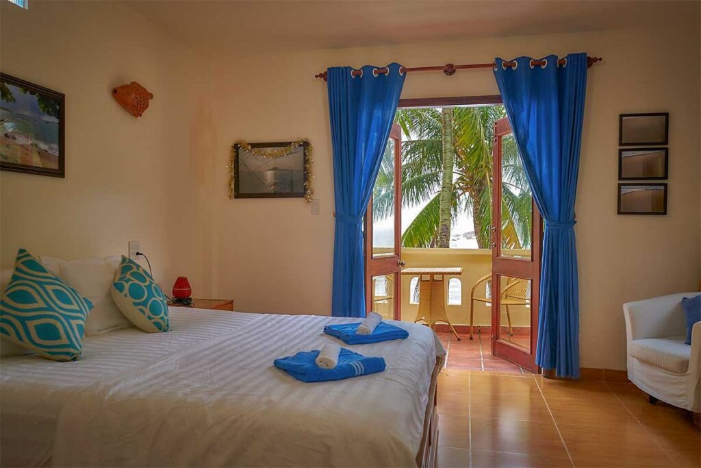 Guestroom at Haven Vietnam on Bai Xep Beach – Cozy room with sea-view balcony, blue curtains, and palm trees outside, offering a simple but beachfront stay.