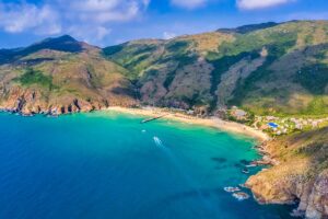 Drone view of Ky Co Beach, Quy Nhon, with turquoise waters, a jetty, sunbeds, and mountain backdrop along the Phuong Mai Peninsula.