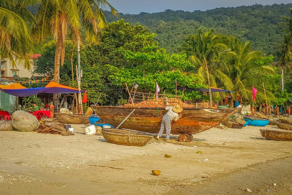 Woman maintaining a wooden fishing boat on the beach of Cham Island, surrounded by palm trees and coracle boats.