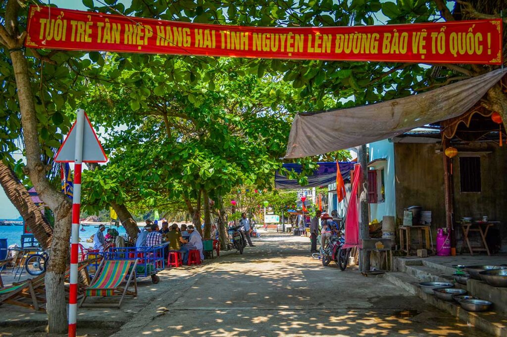 Quiet street in Cham Island village with locals relaxing under shade trees and small cafés facing the waterfront.