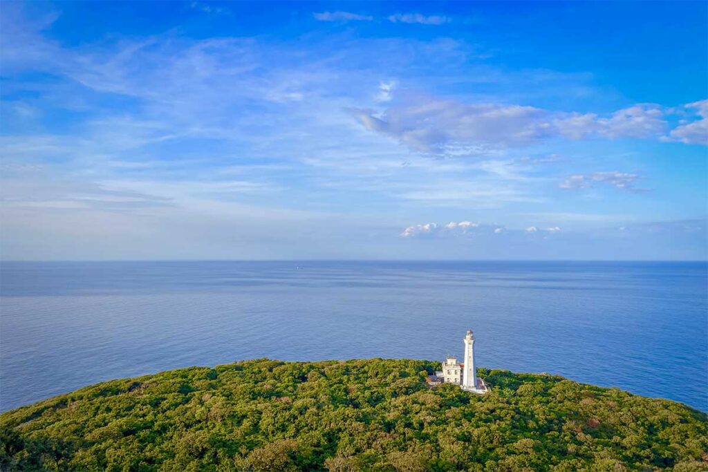 Aerial view of Cham Island lighthouse rising above lush green forest, with endless blue sea stretching to the horizon in central Vietnam.