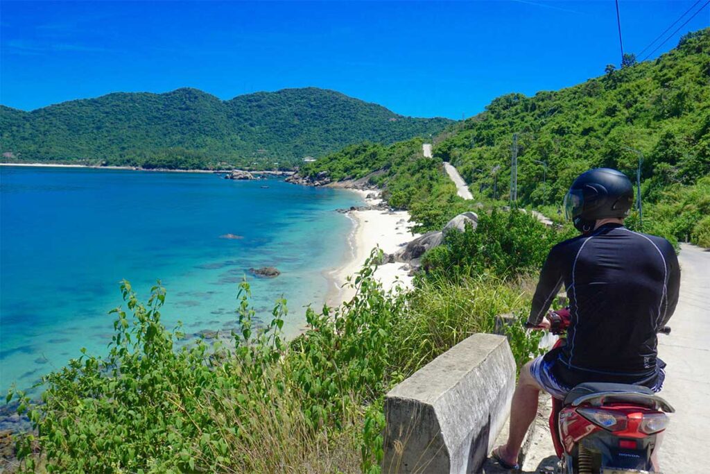 Traveler riding a motorbike along Cham Island’s coastal road, overlooking turquoise water and quiet white-sand beaches.