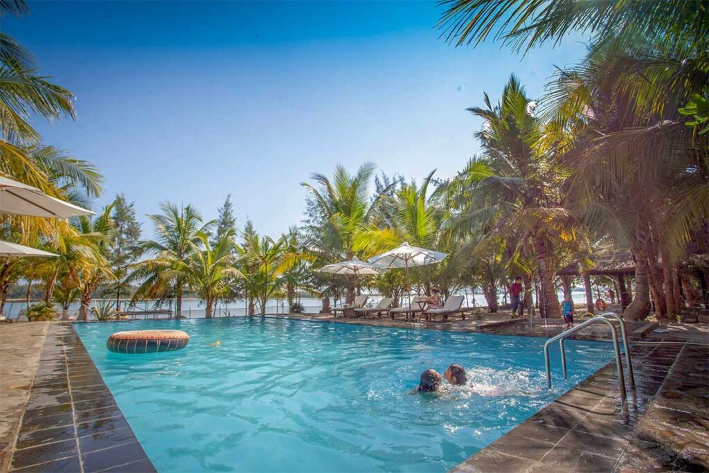 Le Domaine de Tam Hai Resort’s poolside area with sun loungers and umbrellas, overlooking the tranquil river and lush greenery.