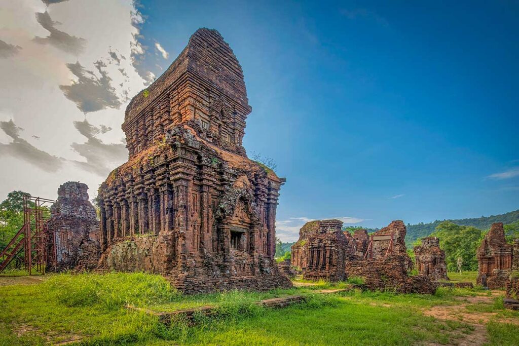 Weathered Cham brick tower under stabilization scaffolding at My Son Sanctuary, set in a grassy valley ringed by low mountains