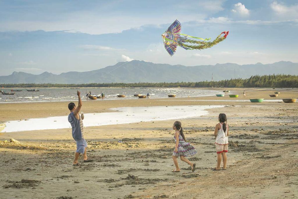A father and children fly a colorful kite on the quiet beach of Tam Hai Island, with round fishing boats and distant mountains in view.