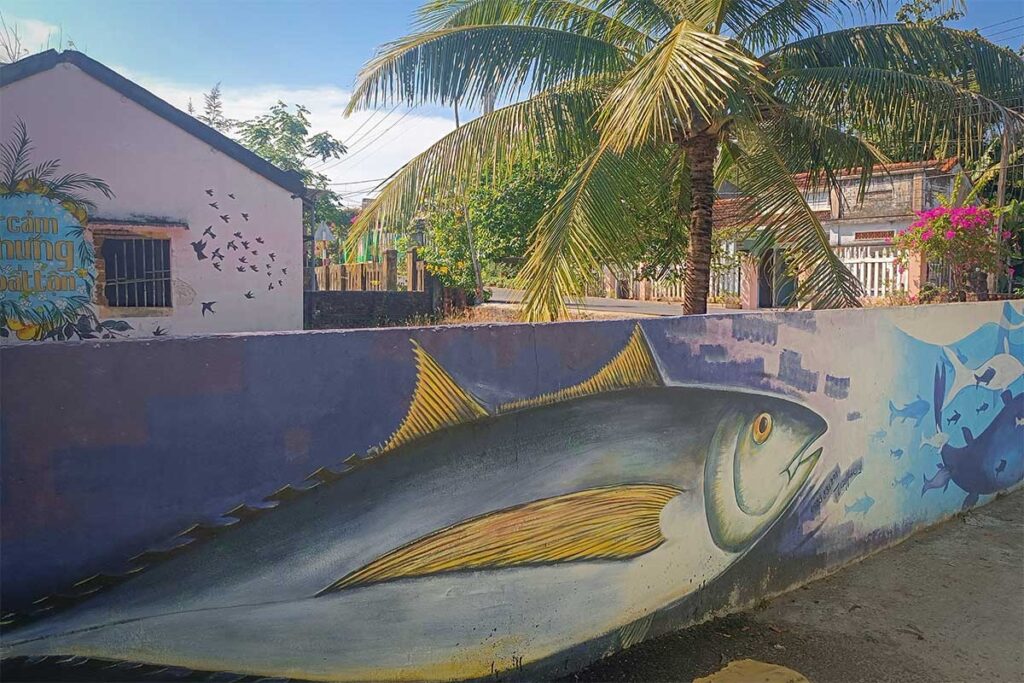 Wall mural of a large fish and flying birds in the mural village on Tam Hai Island, surrounded by palm trees and quiet village streets.