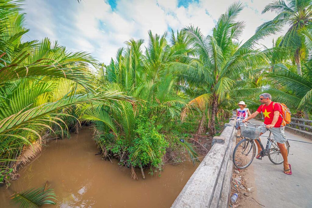 Travelers with bicycles stopping on a small bridge in Ben Tre, Vietnam, to enjoy views of quiet canals and coconut trees.