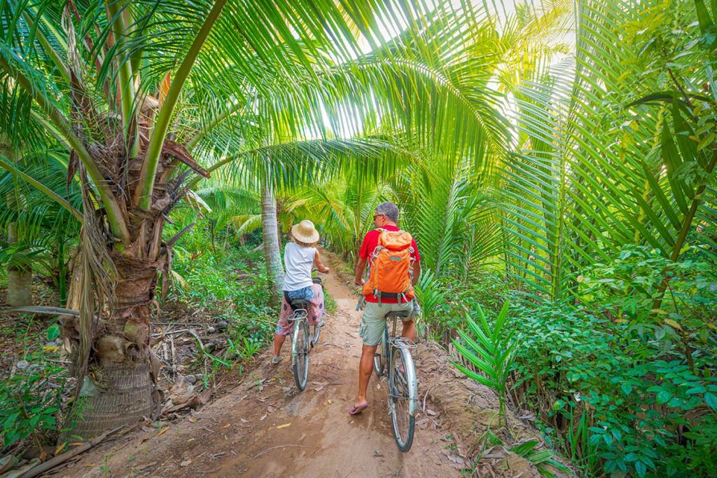 Cyclists exploring rural dirt paths lined with coconut palms in Ben Tre, Vietnam – a highlight of eco-tourism in the Mekong Delta.