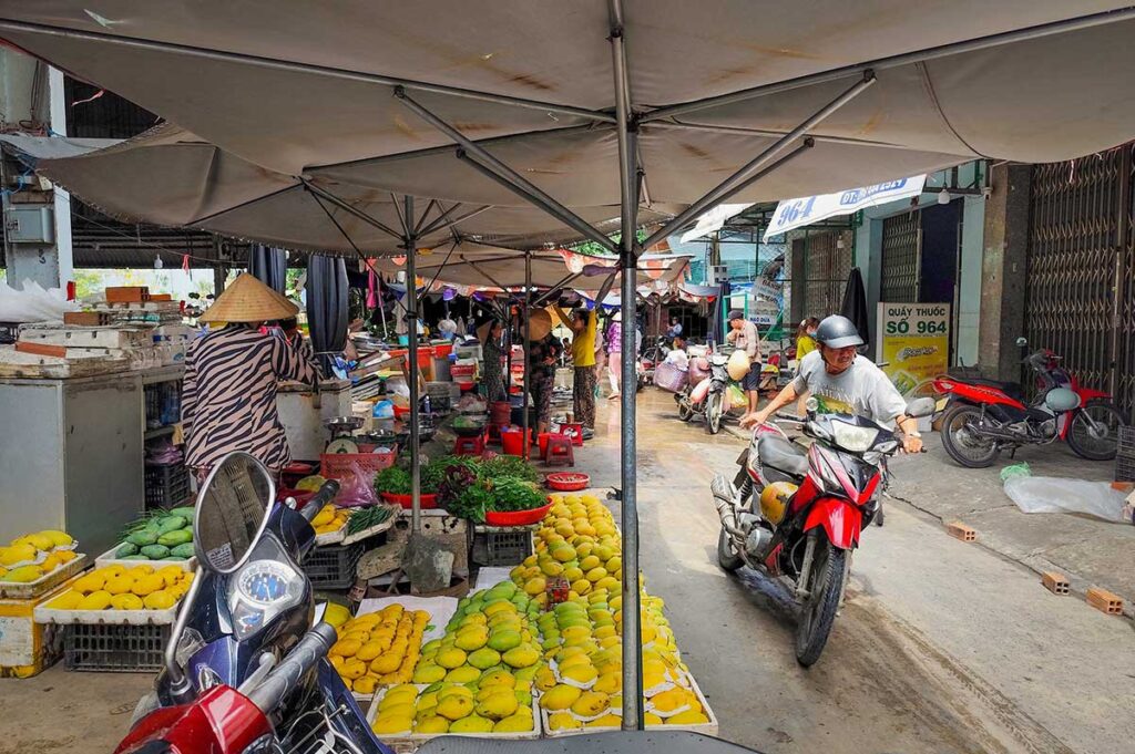 Fresh fruit and mango stalls at Ben Tre Market – Street market scene with colorful produce and motorbikes passing through.