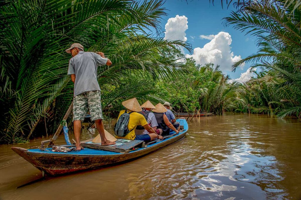 Hand-rowed sampan boat ride in Ben Tre – Local boatman navigating tourists with conical hats through shaded canals.