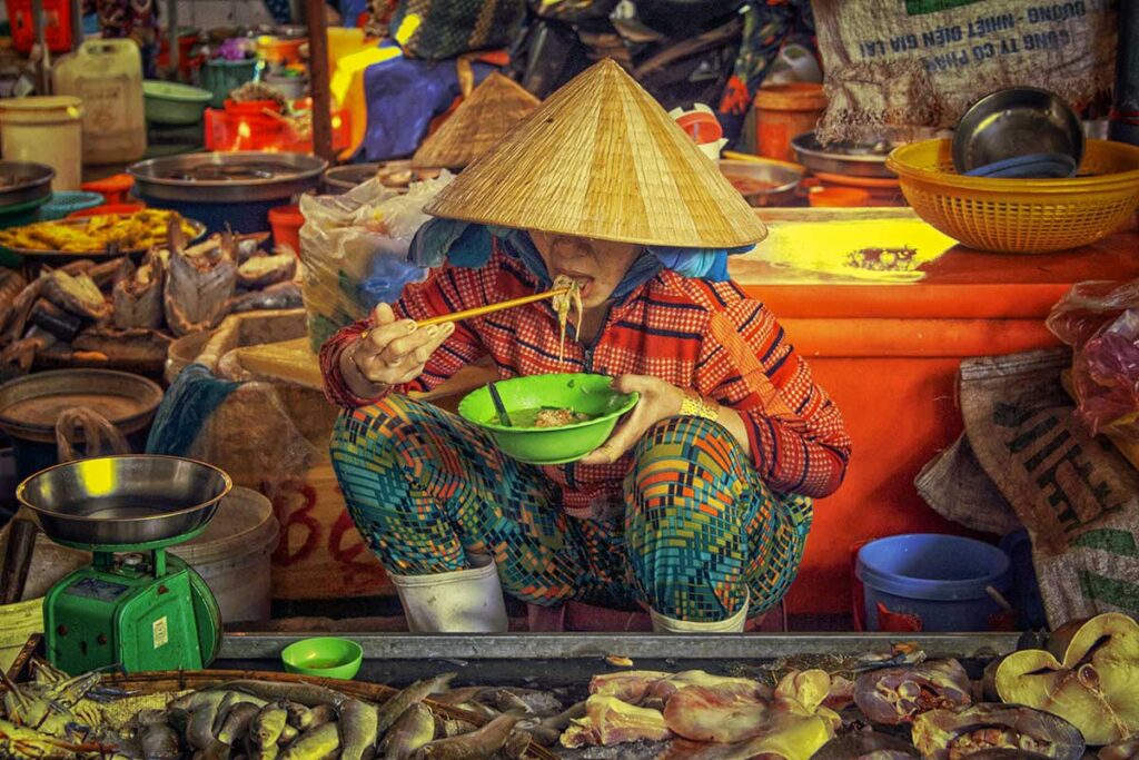 Woman in conical hat eating noodles inside Ha Tien market fish section.