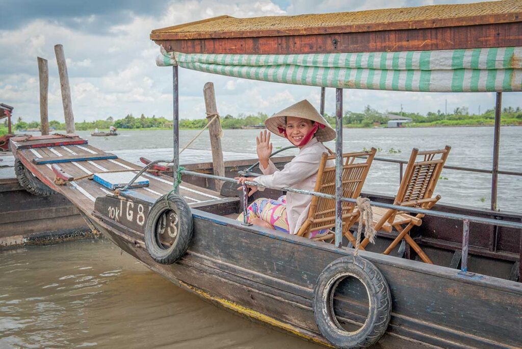 Local woman waving from a boat in Vinh Long – Friendly boat operator greeting visitors on a wooden riverboat tour in the Mekong Delta.