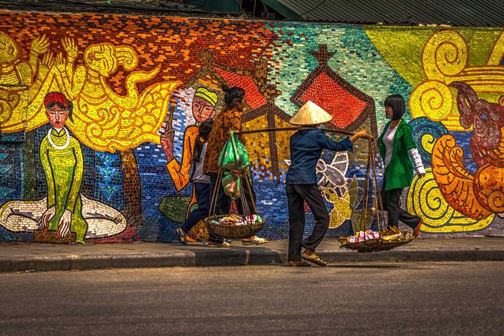 Street Vendor in Front of Hanoi Ceramic Mosaic Mural: A woman carrying a shoulder pole laden with baskets of goods walks in front of the colorful Hanoi Ceramic Mosaic Mural.