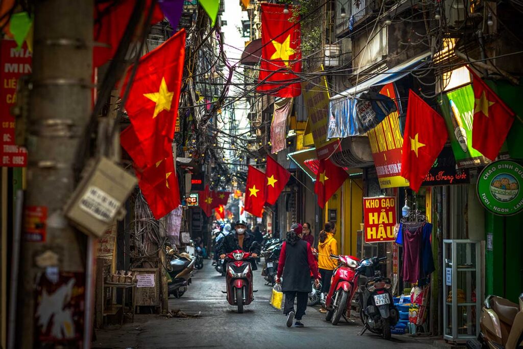 A bustling street scene in Hanoi, a vibrant city in Vietnam, with motorbikes zipping through narrow lanes, locals walking past colorful shophouses adorned with Vietnamese flags.