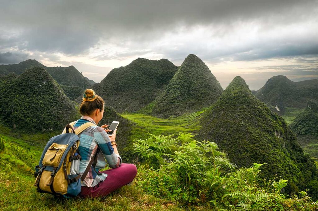 Woman sitting alone in the grass, watching her phone, with stunning mountain views in Ha Giang, Vietnam.