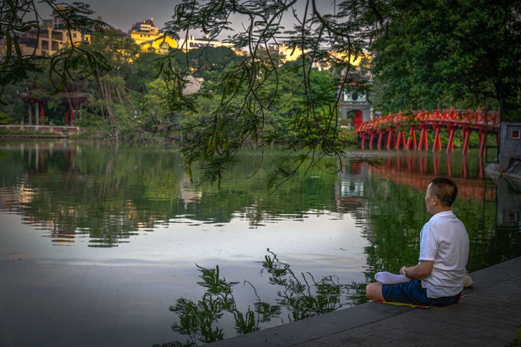 Hanoi in the morning at Hoan Kiem Lake - A man sitting next to the lake meditating with the red Huc Bridge in the background