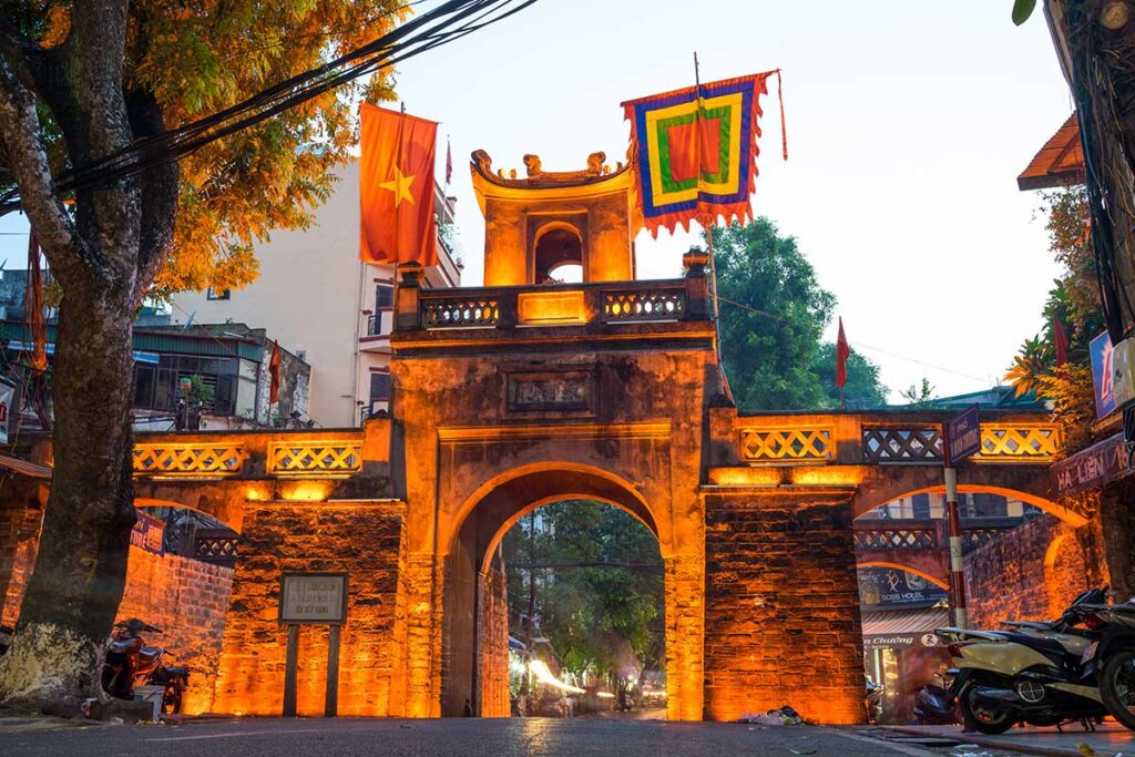Hanoi Old City Gate (O Quan Chuong) softly illuminated at night, with motorbikes passing through and street activity around the arches.