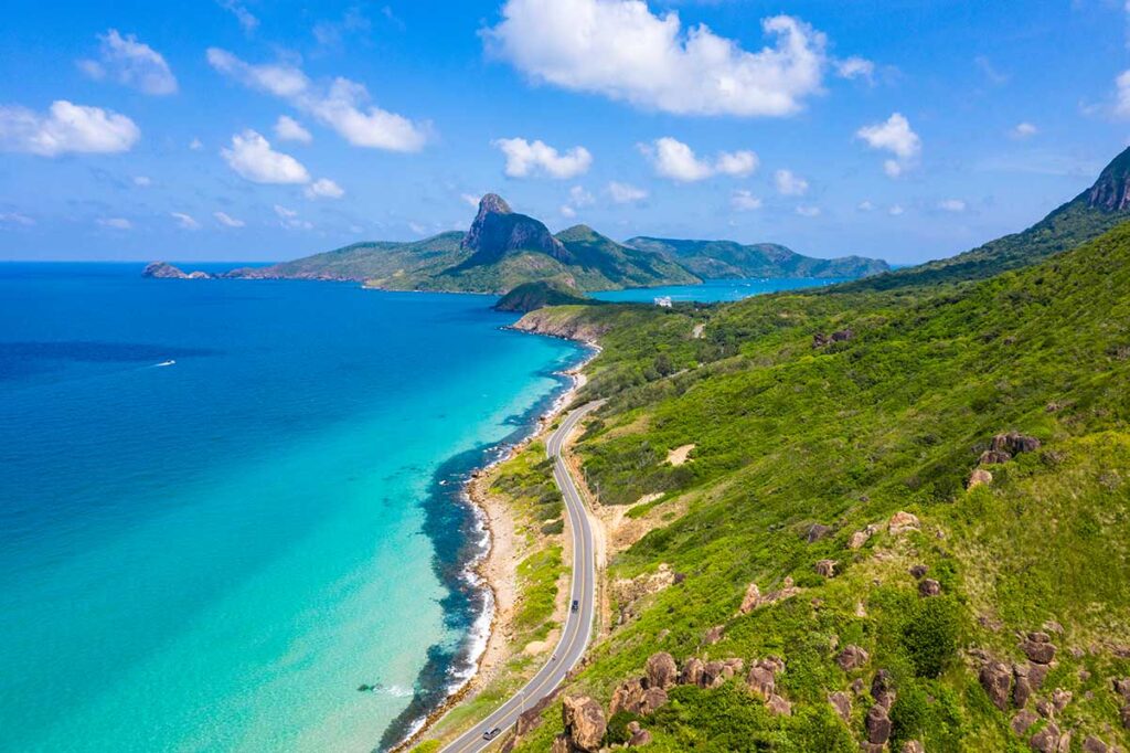 Things to do in Con Dao – A stunning aerial view of a scenic coastal road, winding through lush green mountains with islands and the turquoise ocean in the background, showcasing the island’s breathtaking landscapes and perfect conditions for motorbike exploration.