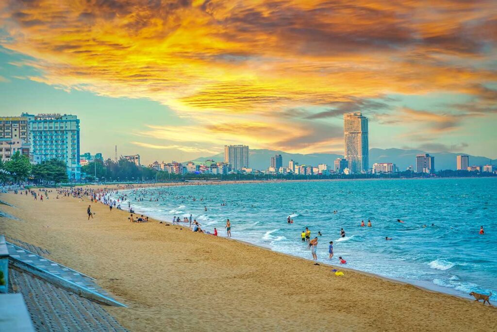 Sunset over Quy Nhon City Beach with locals swimming and walking along the golden sand, backed by high-rise hotels and a vibrant seafront skyline.
