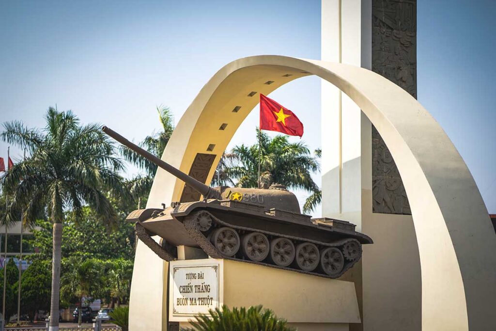 A close-up photograph of a tank, part of the Buon Ma Thuot Victory Monument, displaying the Vietnamese flag atop.