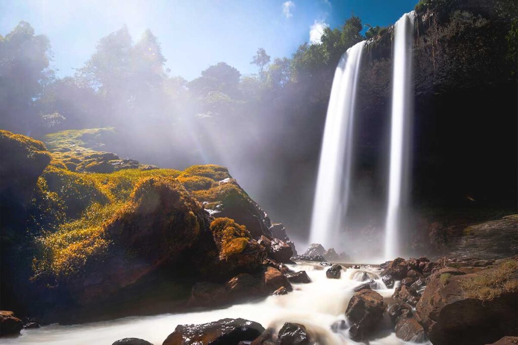 Sunlight filtering through the trees, casting beams of light onto the rocks and flowing water at Dak G’Lun Waterfall.