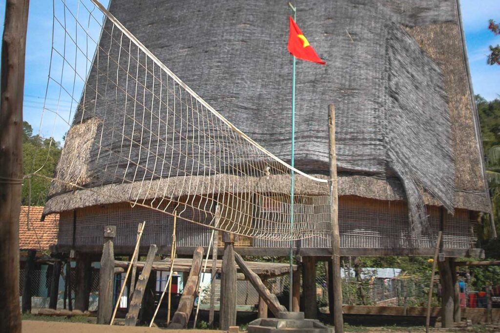 A tall-roofed stilt house in an ethnic village, with a volleyball court and a Vietnamese flag in the foreground, resembling the architecture found in Kon Tum.