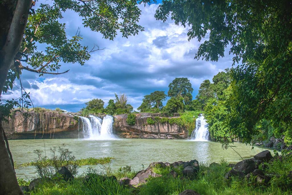 A peaceful path surrounded by lush trees, leading to Dray Sap Waterfall, where two streams of water cascade into a natural pool.