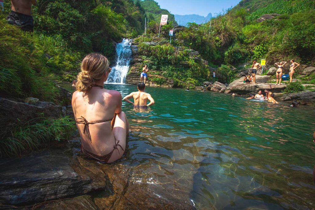 A girl sitting in a bikini on a rock, watching people swim and jump from the rocks at Du Gia Waterfall, a