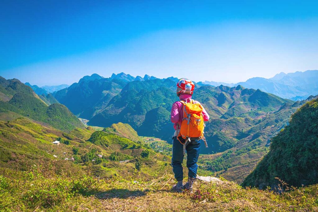 Ha Giang Loop in April – A female traveler wearing a motorbike helmet stands on a mountain viewpoint, admiring the stunning blue-sky scenery of Ha Giang Loop in April.