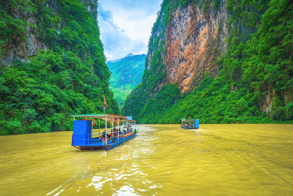 A tourist boat cruising along the brown waters of the Nho Que River, passing between the towering cliffs of Ma Pi Leng Pass in Ha Giang.