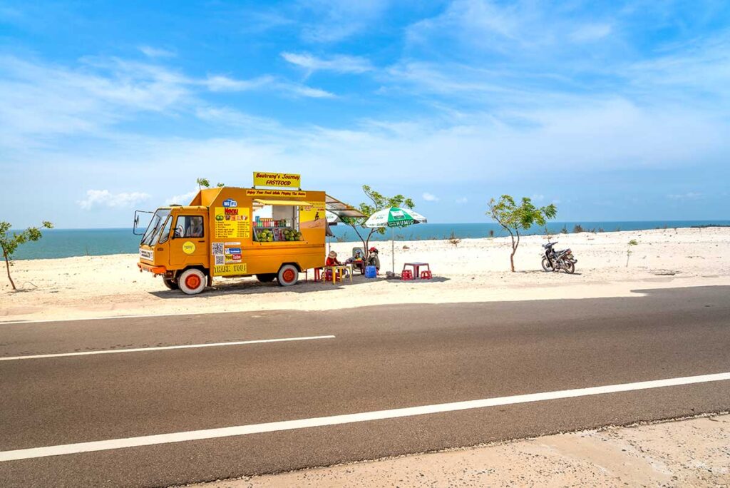 Mui Ne in April: A small fruit truck parked along the scenic coastline of Mui Ne in April, with a stunning blue sky and light clouds creating a picturesque roadside view.