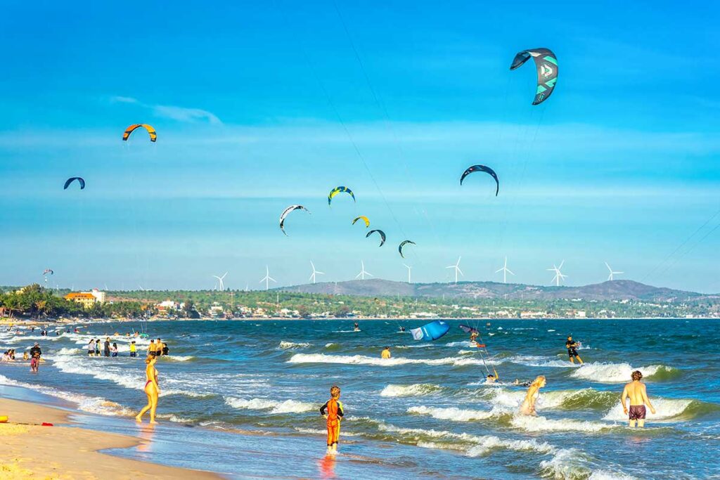 Mui Ne in February: A vibrant scene of Mui Ne in February, with numerous kite surfers riding the strong winds over the turquoise sea, capturing the peak of kite surfing season.