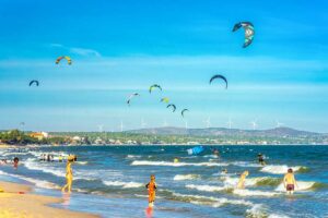 Mui Ne in February: A vibrant scene of Mui Ne in February, with numerous kite surfers riding the strong winds over the turquoise sea, capturing the peak of kite surfing season.