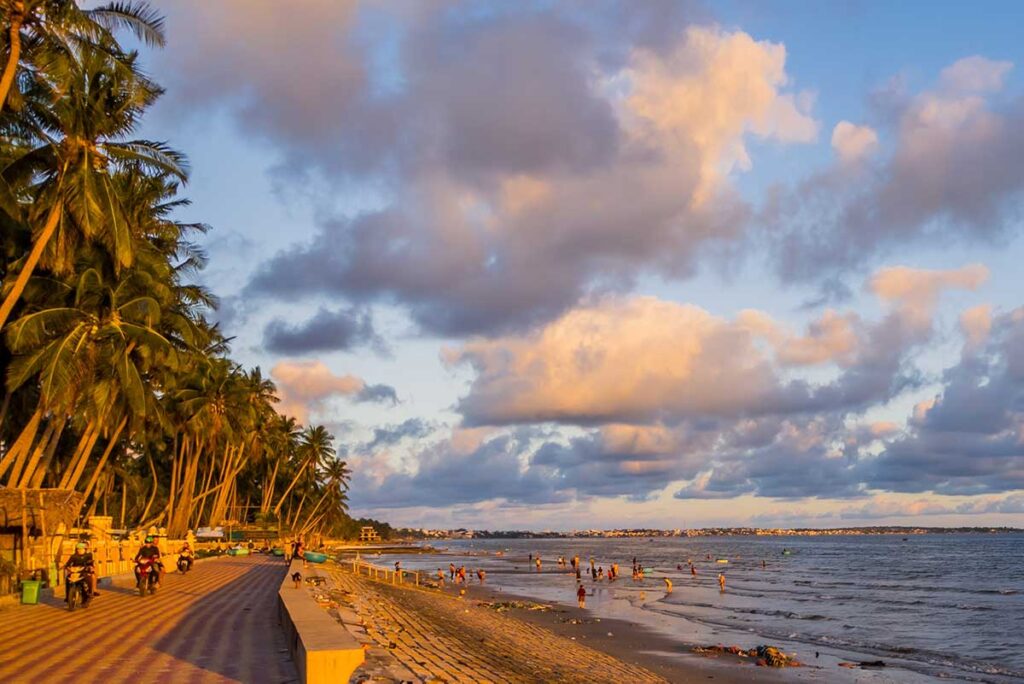 Mui Ne in October: A concrete beachfront near Mui Ne in October, possibly Phan Thiet, with a cloudy sky and a warm, glowing sunset reflecting off the water.