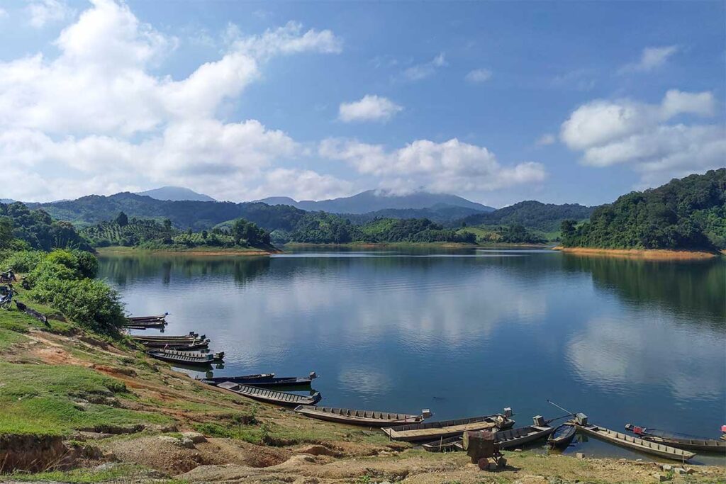 A calm lakeside scene with wooden boats resting on the shore, facing the lush forests of Nam Kar Nature Reserve.