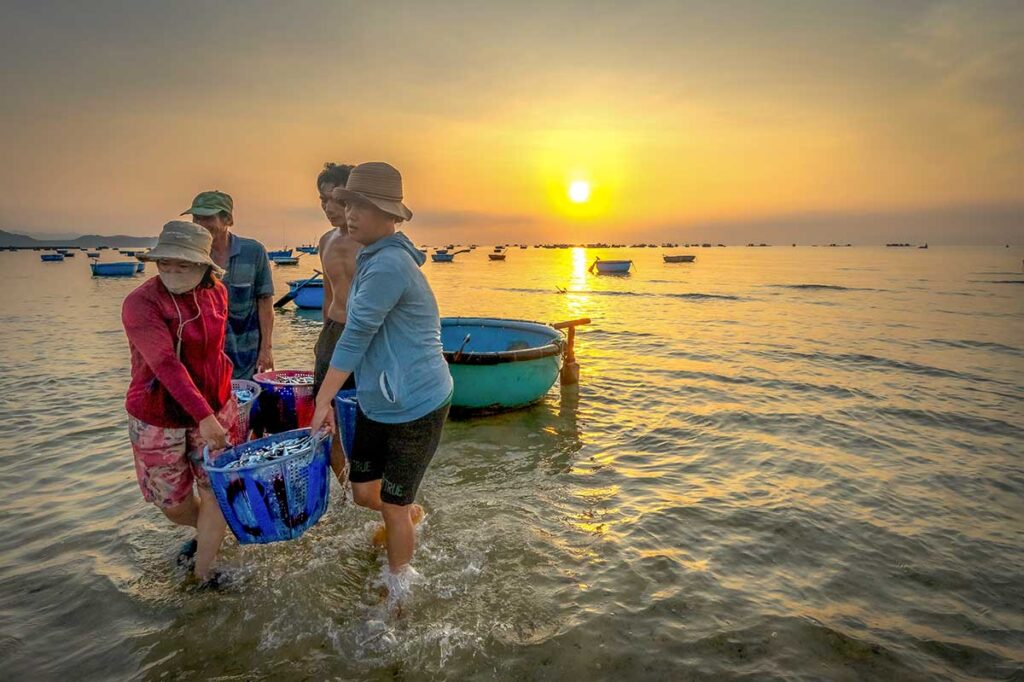 Fishermen carrying baskets of fresh catch from coracle boats at sunrise on Phan Rang’s coast.