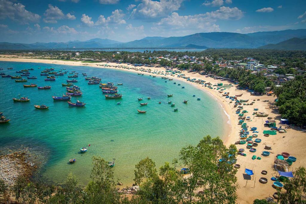 Colorful fishing boats anchored at a traditional beach village in Phu Yen, Vietnam, with golden sand, palm trees, and mountain view
