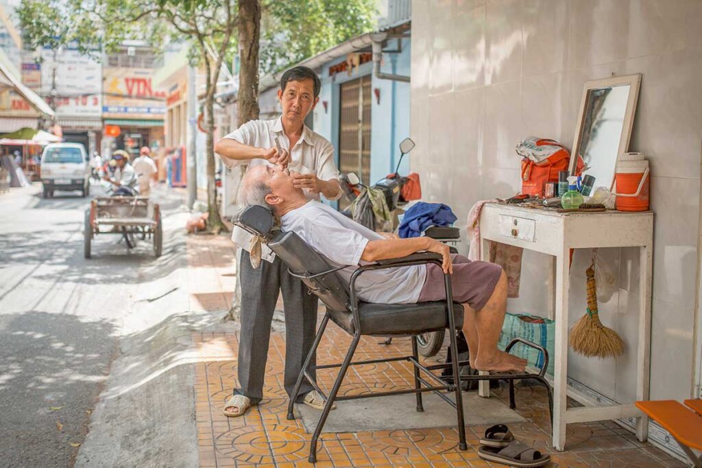 Traditional street barber giving a shave to an elderly man on the sidewalk in Soc Trang’s downtown area.