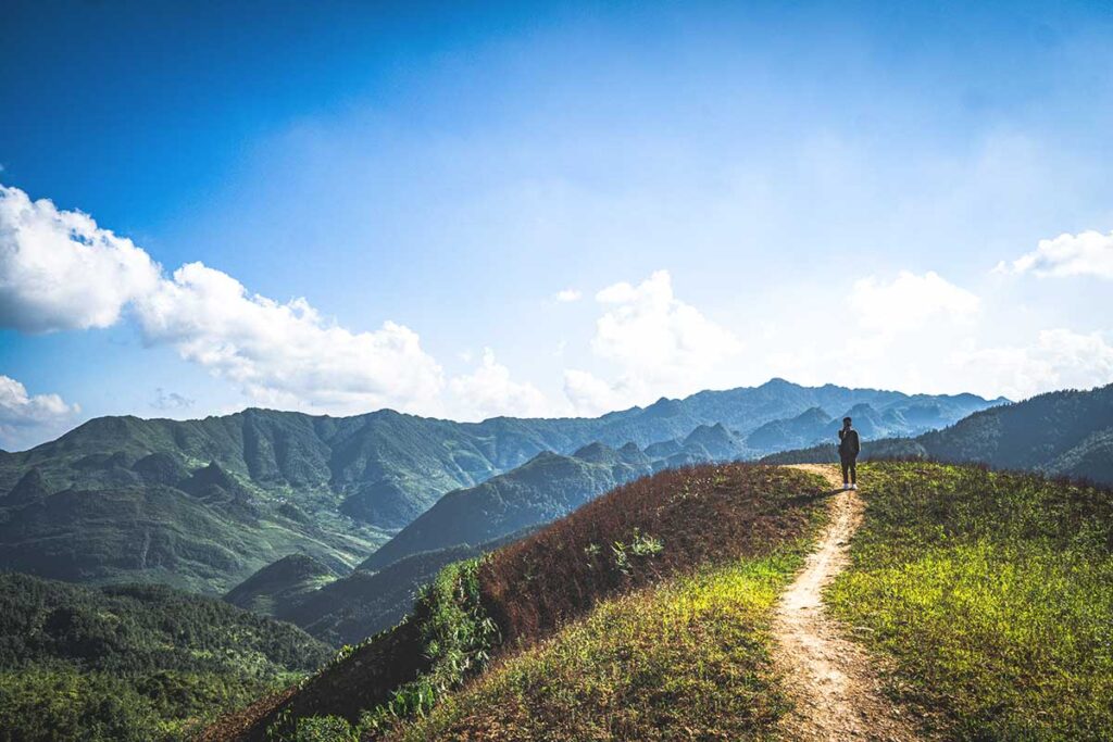 A traveler trekking along a small dirt trail on top of a mountain, enjoying a clear blue sky day with expansive mountain views in Ha Giang.