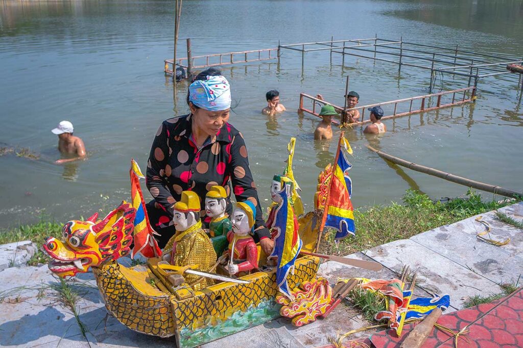 A woman sitting on the edge of the water with water puppets and boat prop with local man setting up a water puppet theater in the water behind her at Water Puppet Village in Thanh Hai Commune