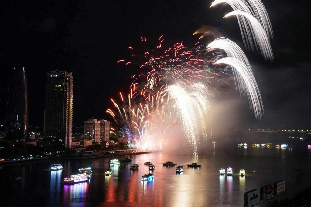 Spectators watching the Da Nang International Fireworks Festival from a river cruise on the Han River, with colorful fireworks reflecting on the water.