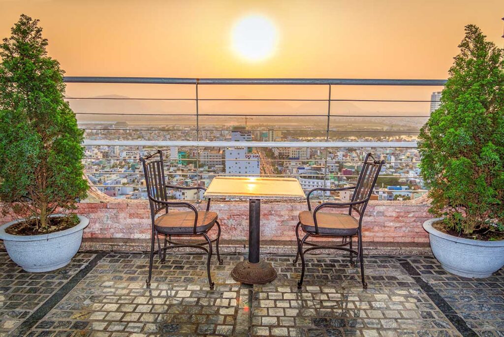 Da Nang in March – A rooftop bar setting with two chairs and a table, overlooking the city skyline during a breathtaking sunset over Da Nang.