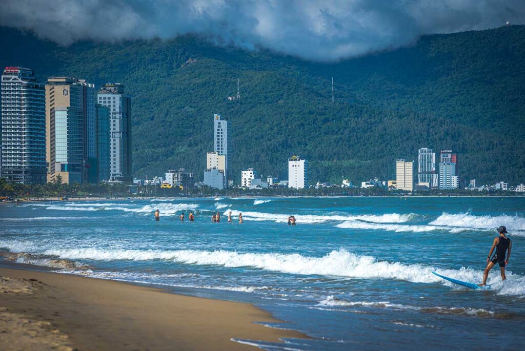 Da Nang in October – A surfer riding the waves, with other beachgoers enjoying the ocean, marking the surf season in Da Nang.