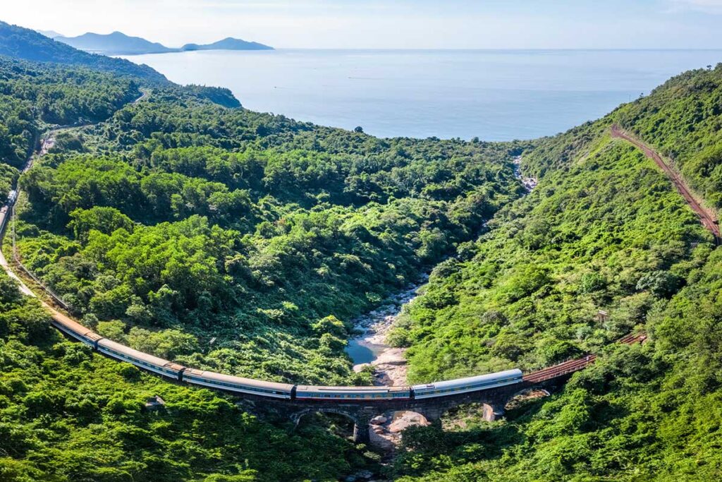 A drone shot of a train crossing a railway bridge along the Hai Van Pass, with the coastline stretching into the background.