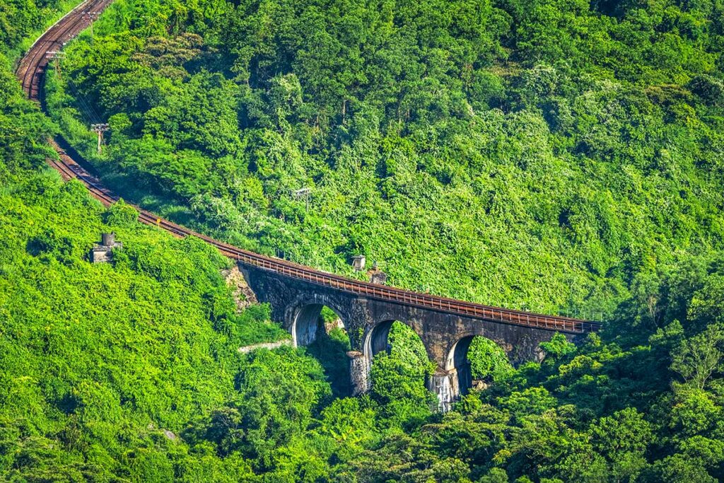 A high-angle drone shot of the railway bridge along the Hai Van Pass, surrounded by lush greenery and the coastal waters.