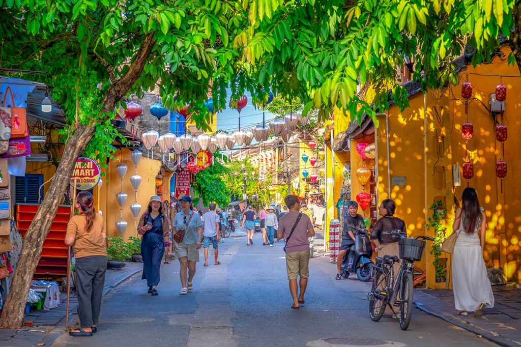 Visitors walking through lantern-filled streets of Hoi An Ancient Town in July, enjoying the lively atmosphere despite the midday heat and occasional short showers.