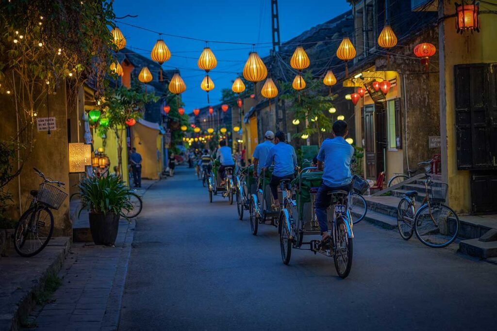 Cyclo drivers riding through lantern-lit streets of Hoi An during the Lantern Festival, showcasing the town’s romantic evening glow.