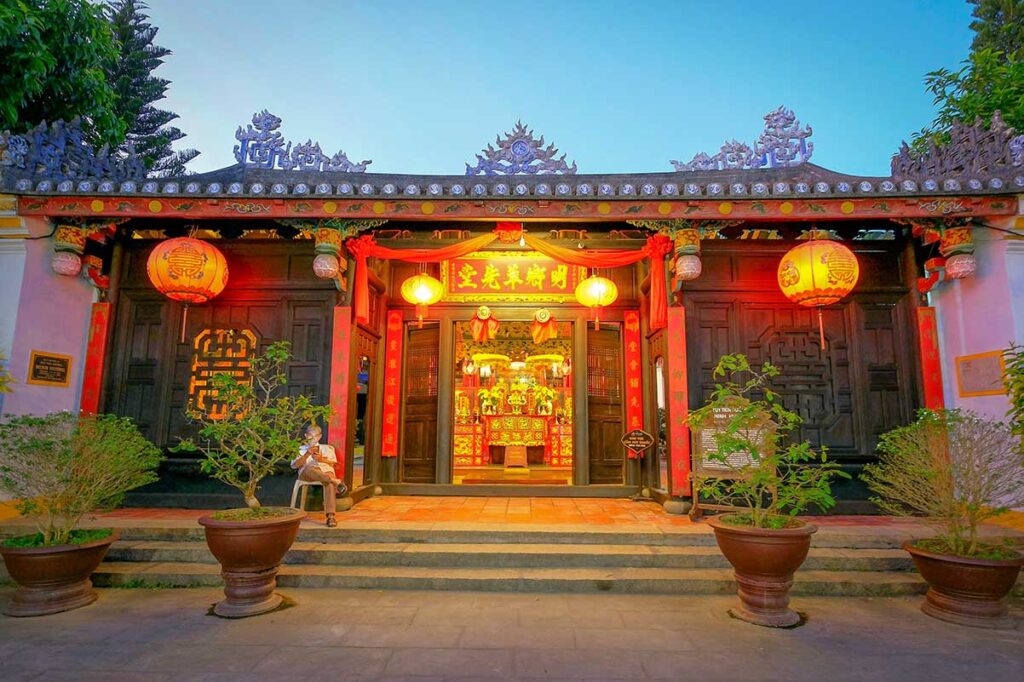 Visitors stopping at a brightly lit temple during the Hoi An Lantern Festival, with red lanterns and traditional Chinese-Vietnamese architecture glowing at dusk.